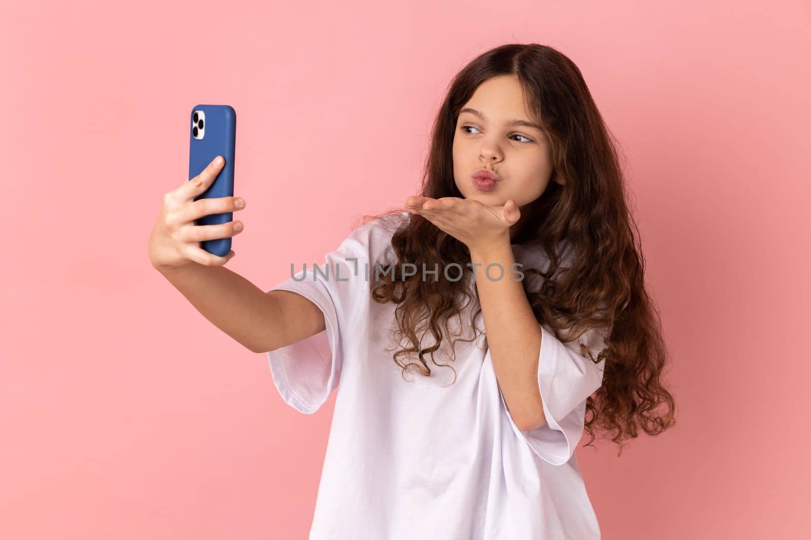 Portrait of little girl wearing white T-shirt sending air kiss on smartphone camera while communicating by video call or streaming vlog. Indoor studio shot isolated on pink background.