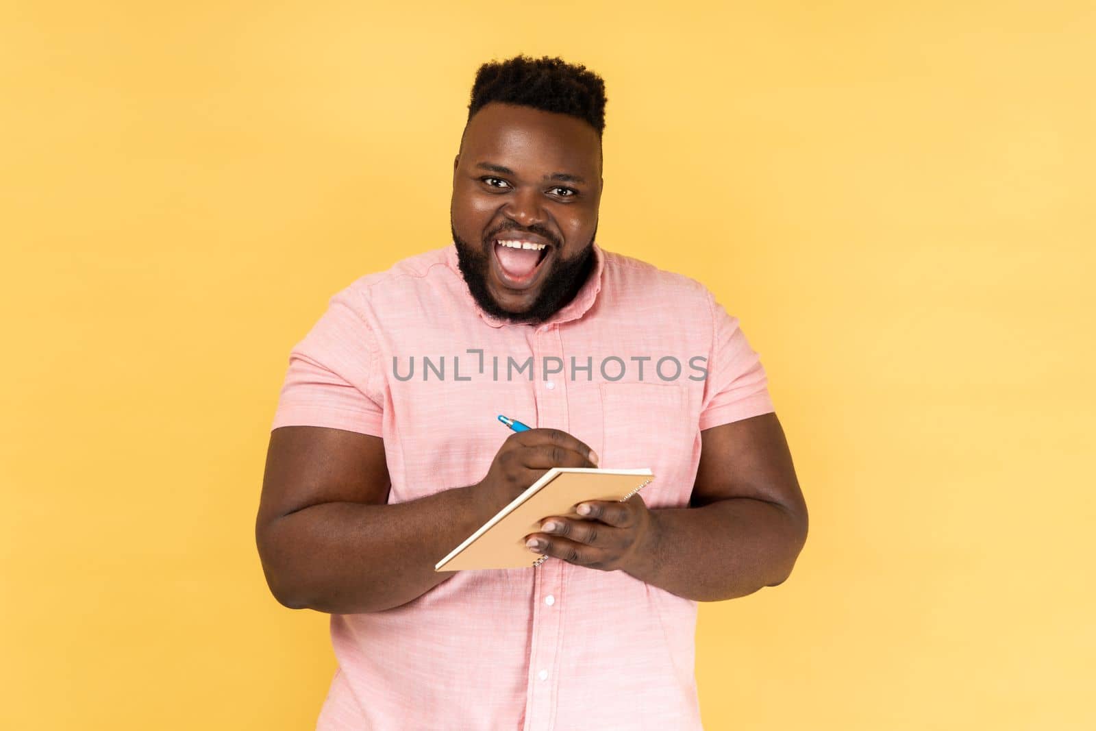 Portrait of man wearing pink shirt writing down in paper notebook, making to do list, having good mood, looking at camera with toothy smile. Indoor studio shot isolated on yellow background.