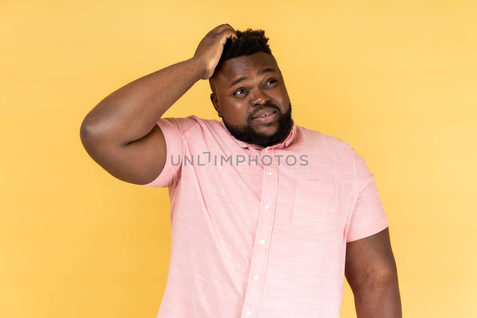 Let me think. Portrait of pensive man wearing pink shirt holding hand on head and thinking, pondering idea, looking away with thoughtful expression. Indoor studio shot isolated on yellow background.