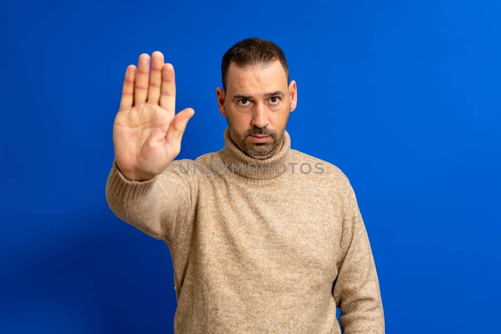 Hispanic handsome man with beard wearing a beige turtleneck sweater over isolated blue background doing stop gesture