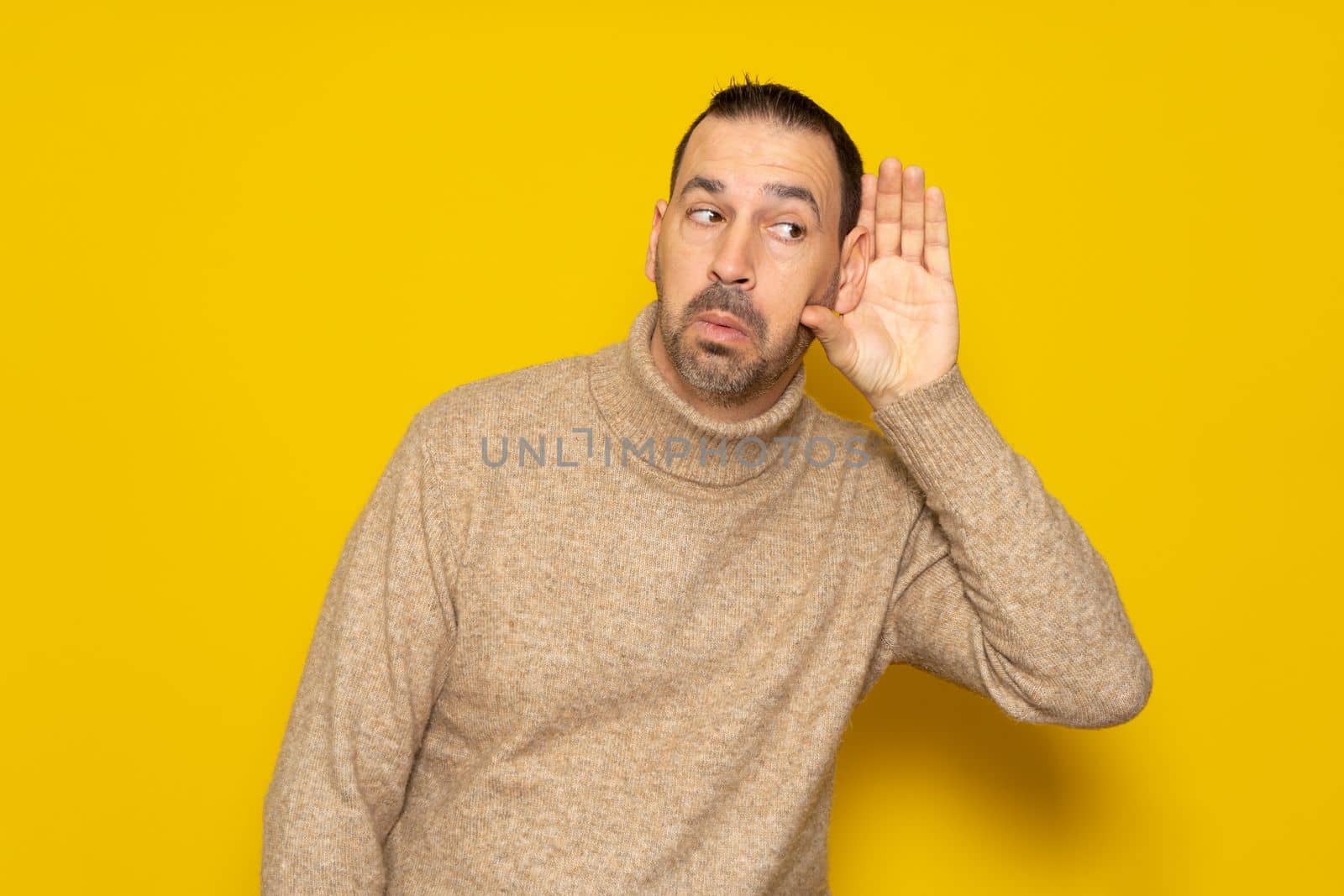 Portrait of a latino man dressed in a beige turtleneck posing isolated over a yellow background turning his ear listening and listening to rumors or gossip. Deafness concept.
