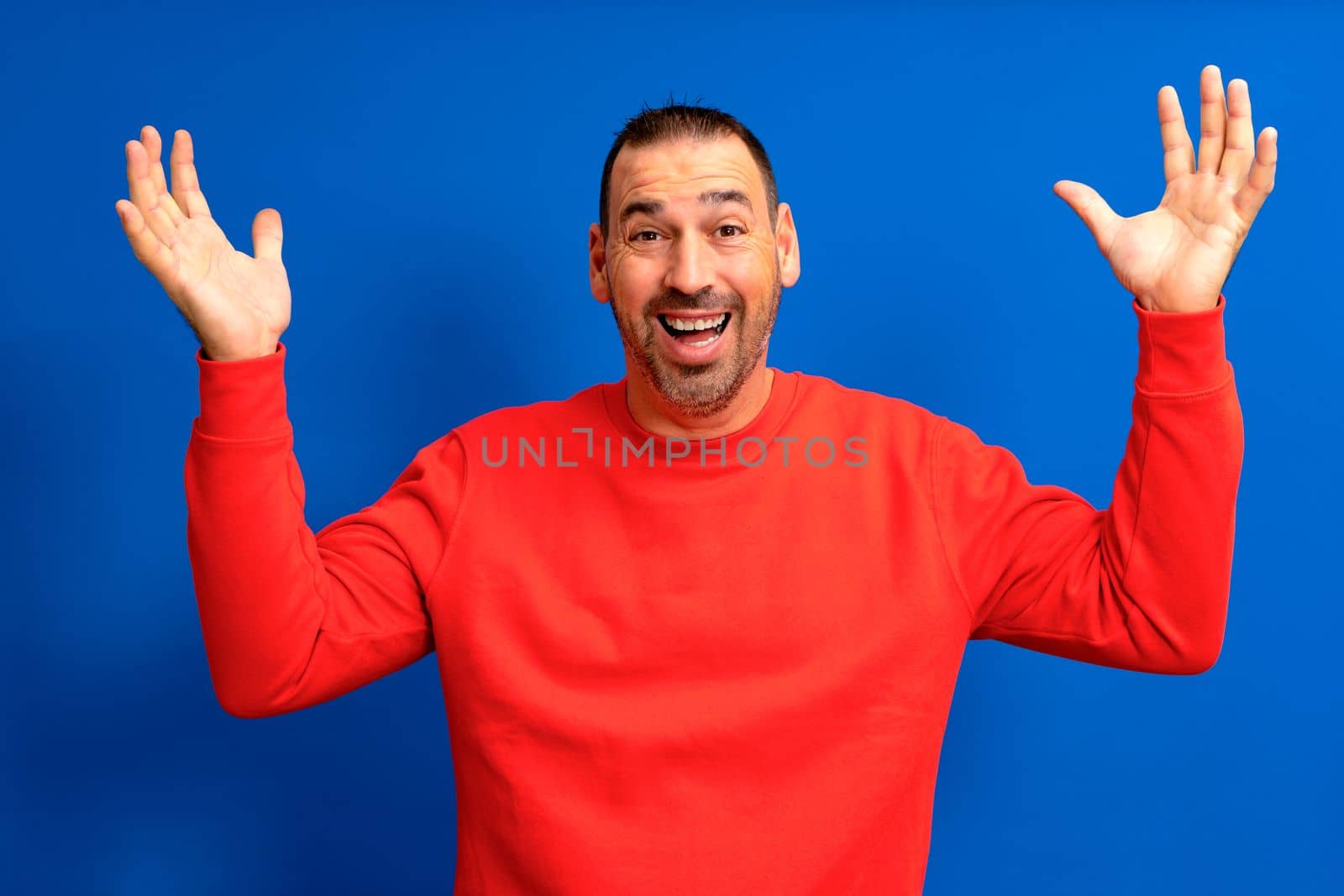 Dreamy bearded melancholic european caucasian man surprised, attractive man 40s in red pullover spreading hands isolated on blue color background studio portrait. People lifestyle concept