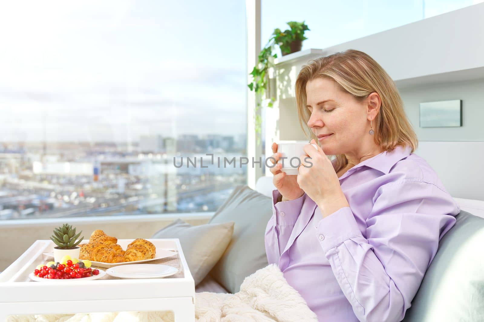 Happy middle aged, beautiful woman having breakfast, relaxing in hotel bed. Woman having breakfast sitting on her bed in luxury apartments. enjoying lazy morning lifestyle