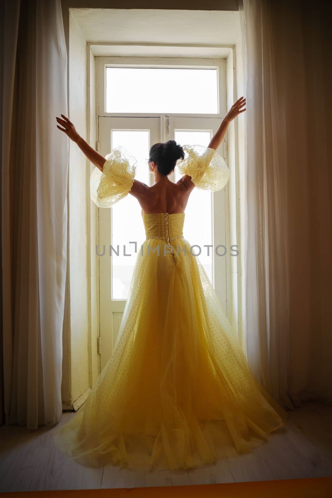 A woman's silhouette in a golden luxurious dress against the background of a window holds a curtain with her hands. Elegant lady in a yellow long silk dress with bare back, back view