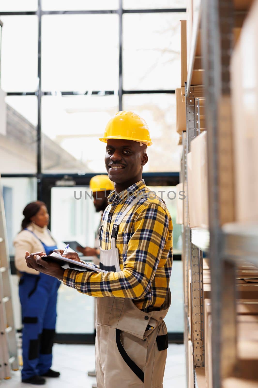 Retail storehouse smiling operator holding clipboard with instructions and looking at camera. African american post office storehouse manager in protective helmet smiling portrait