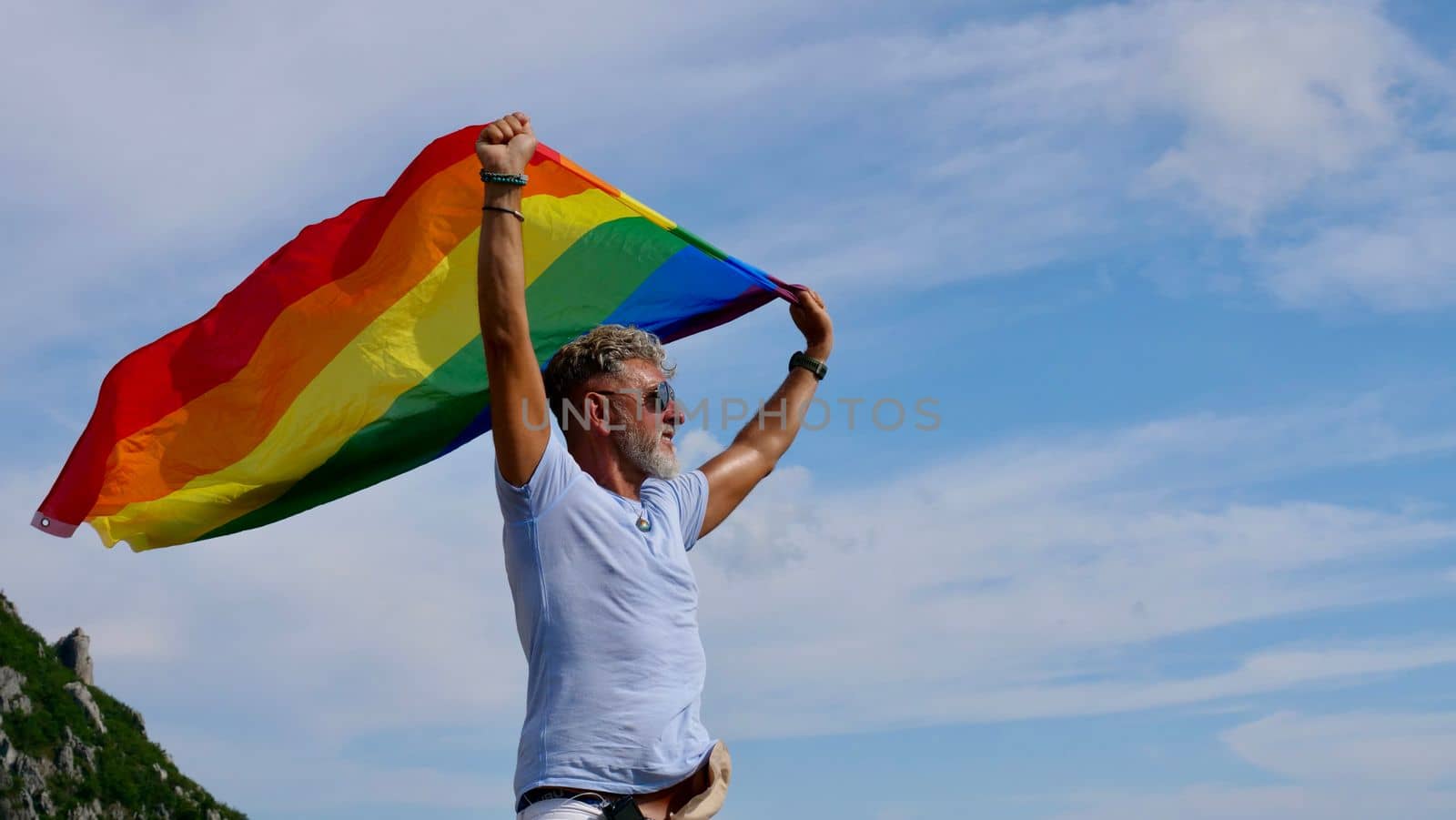 Portrait of a gray-haired elderly Caucasian man with a beard and sunglasses holding a rainbow LGBTQIA flag against a sky background. Bisexual Gay Celebrates Pride Month Coming Out Day