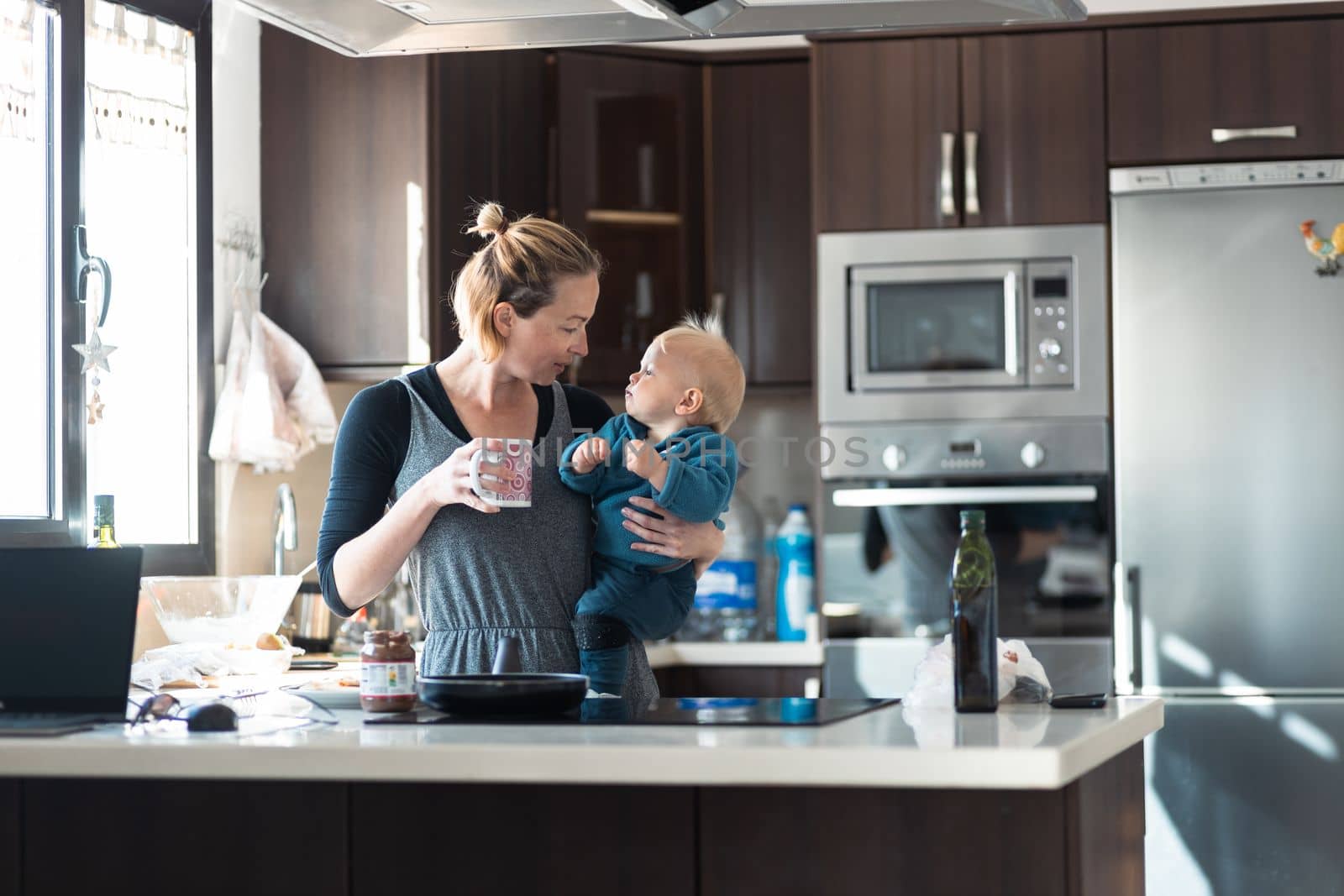 Happy mother and little infant baby boy together making pancakes for breakfast in domestic kitchen. Family, lifestyle, domestic life, food, healthy eating and people concept