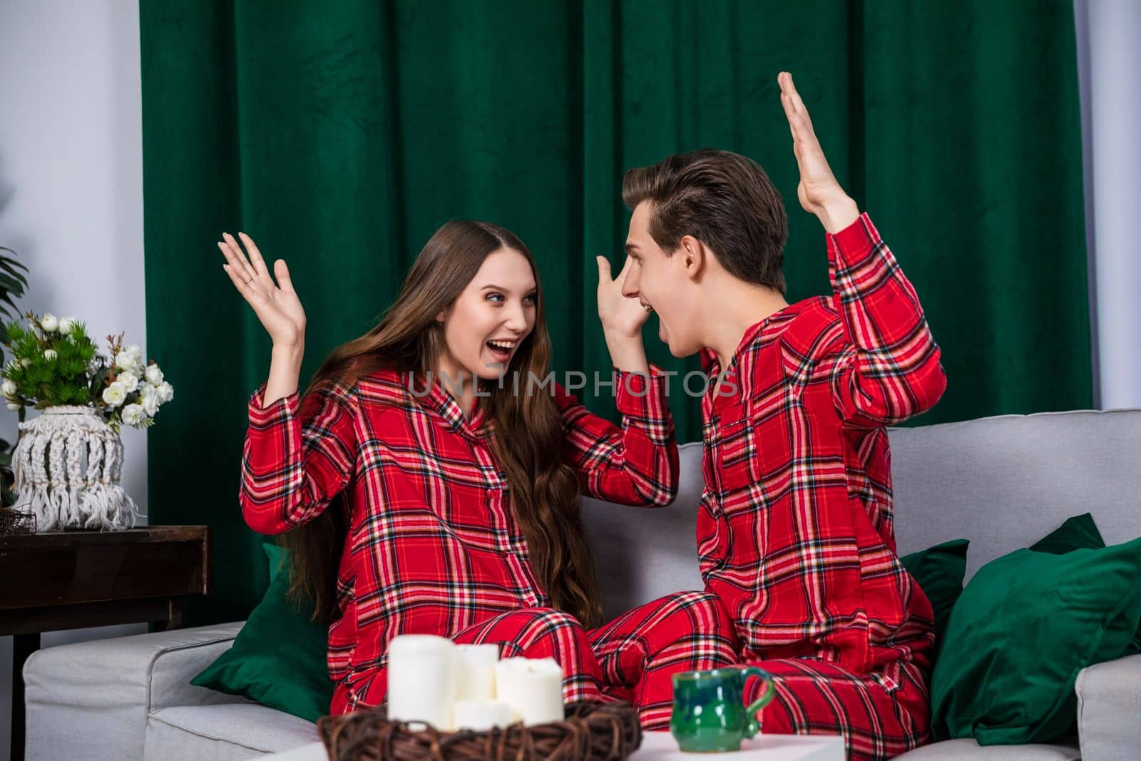 A married couple at a home photo shoot poses in identical pajamas. A happy couple in love spend time sitting on a gray couch. Boy and girl on a date in a romantic interior.