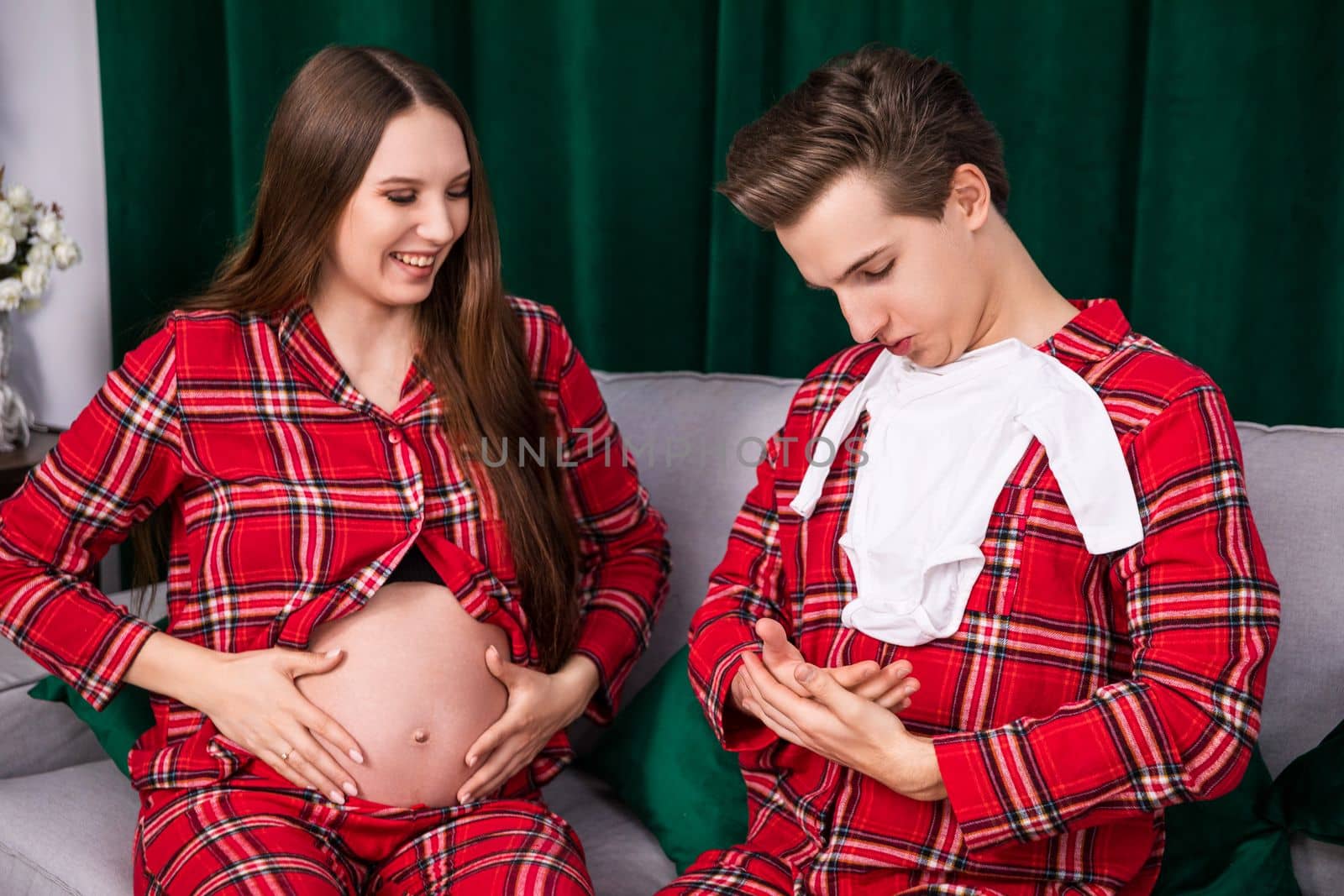 A woman holds her hands over her exposed pregnant belly. A man sitting next to the woman presents infant clothes and arranges his hands as if he is holding a baby on them.