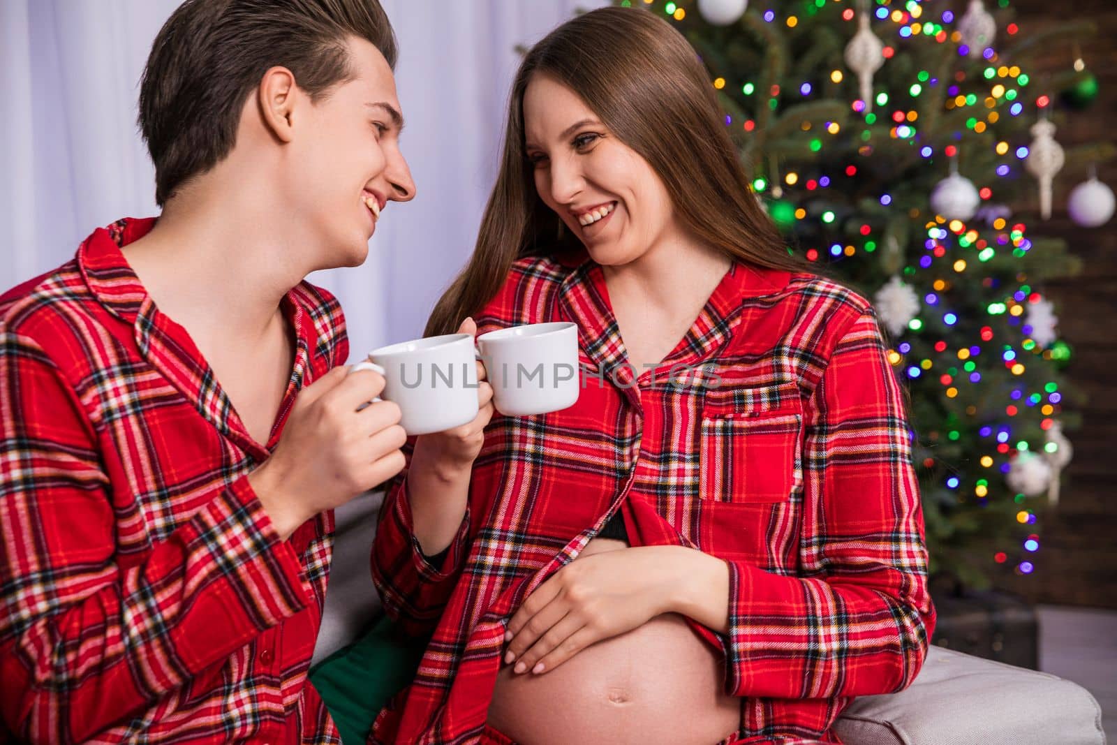 A man and a woman sit close to each other and turn their faces toward each other. The woman is advanced in pregnancy and has an exposed belly. The couple is smiling and holding white cups are toasting.