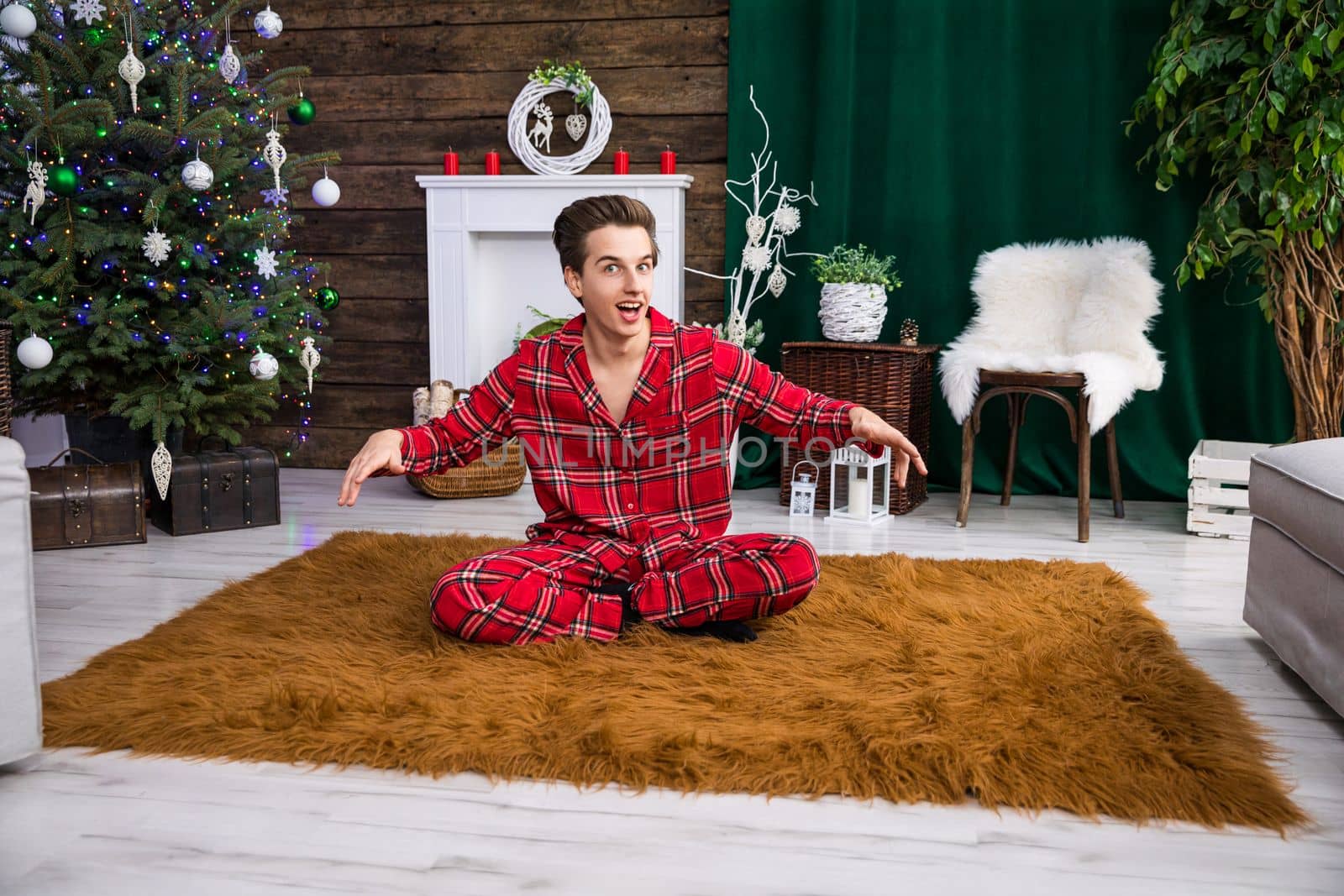 A boy sits on a fluffy brown carpet with his legs crossed and his arms slightly raised. In the background stands a dressed Christmas tree with colorful lights.