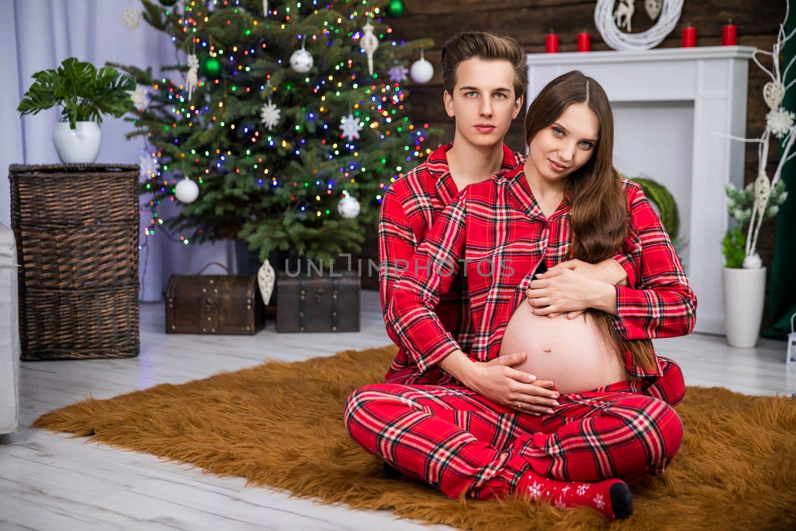 Embracing couple sitting on carpet against background of Christmas tree. The man embraces the pregnant woman from behind. The woman sits with her legs crossed and exposes her pregnant belly.