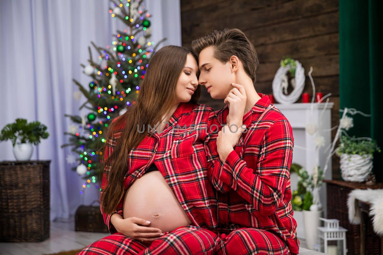 A couple in love show tenderness to each other by hugging and holding hands. The woman sits in front of the man and exposes her pregnant belly. The partners are facing each other.