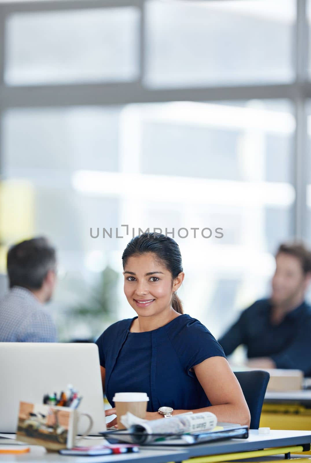 She brings a fresh energy into the office. Portrait of a designer sitting at her desk working on a laptop with colleagues in the background. by YuriArcurs