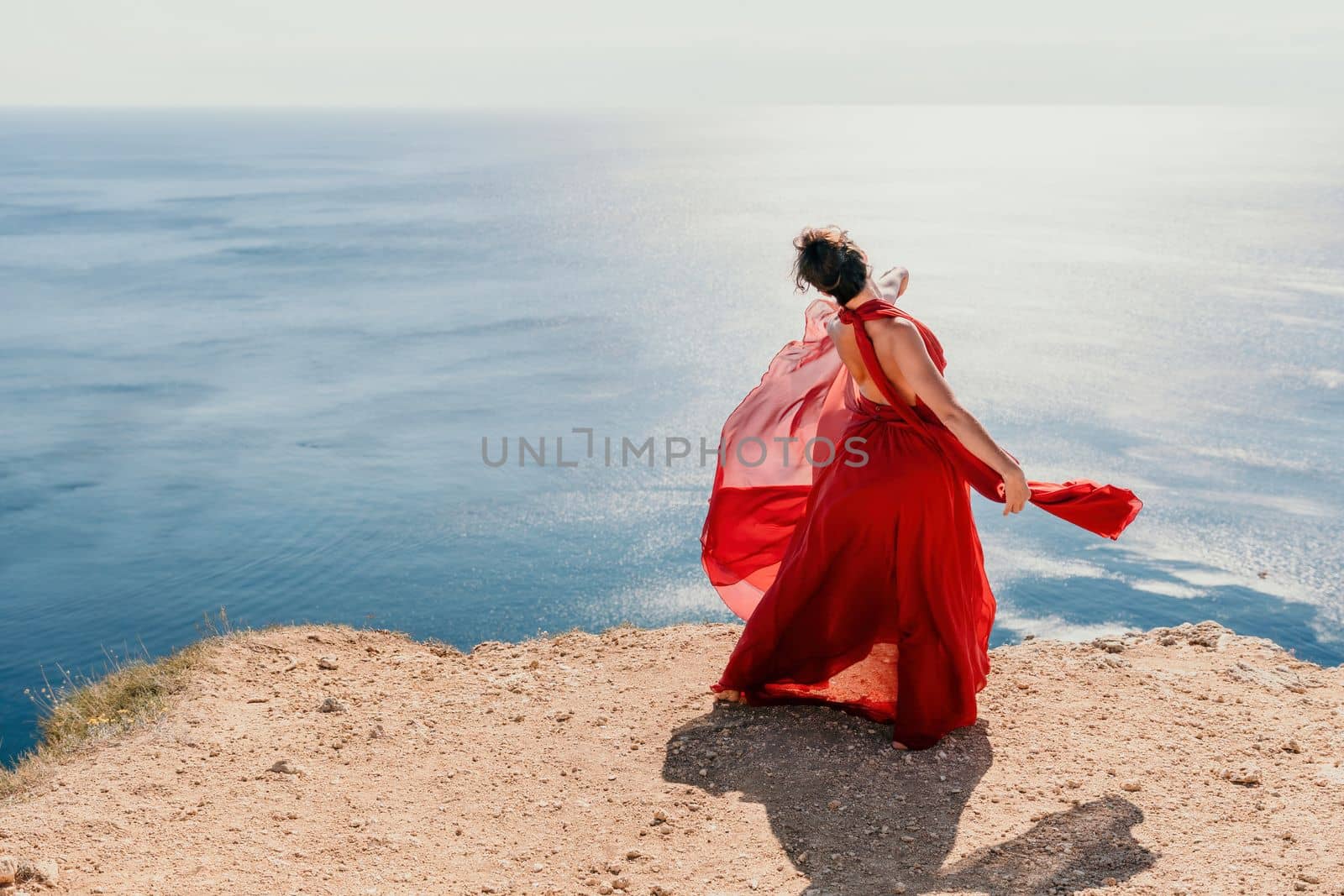 Side view a Young beautiful sensual woman in a red long dress posing on a rock high above the sea during sunrise. Girl on the nature on blue sky background. Fashion photo.
