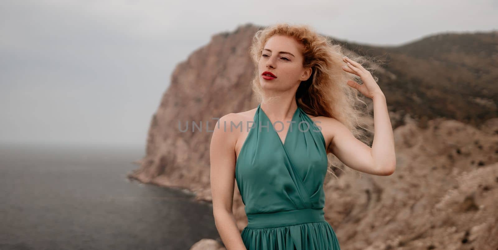 Side view a Young beautiful sensual woman in a mint long dress posing on a volcanic rock high above the sea during sunset. Girl on the nature on overcast sky background. Fashion photo