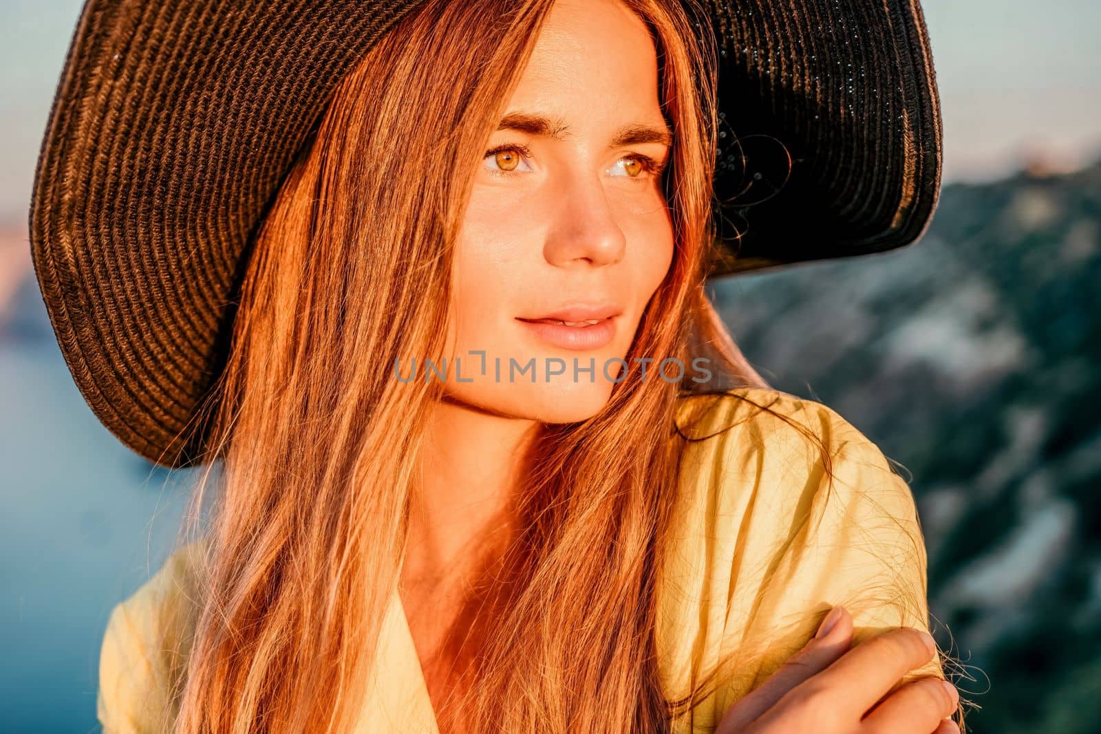 Portrait of happy young woman wearing summer black hat with large brim at beach on sunset. Closeup face of attractive girl with black straw hat. Happy young woman smiling and looking at camera at sea