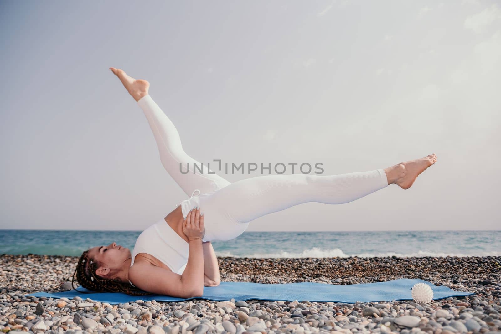 Woman yoga sea. Well looking middle aged woman with braids dreadlocks in white leggings and tops doing stretching pilates on yoga mat near sea. Female fitness yoga routine concept. Healthy lifestyle