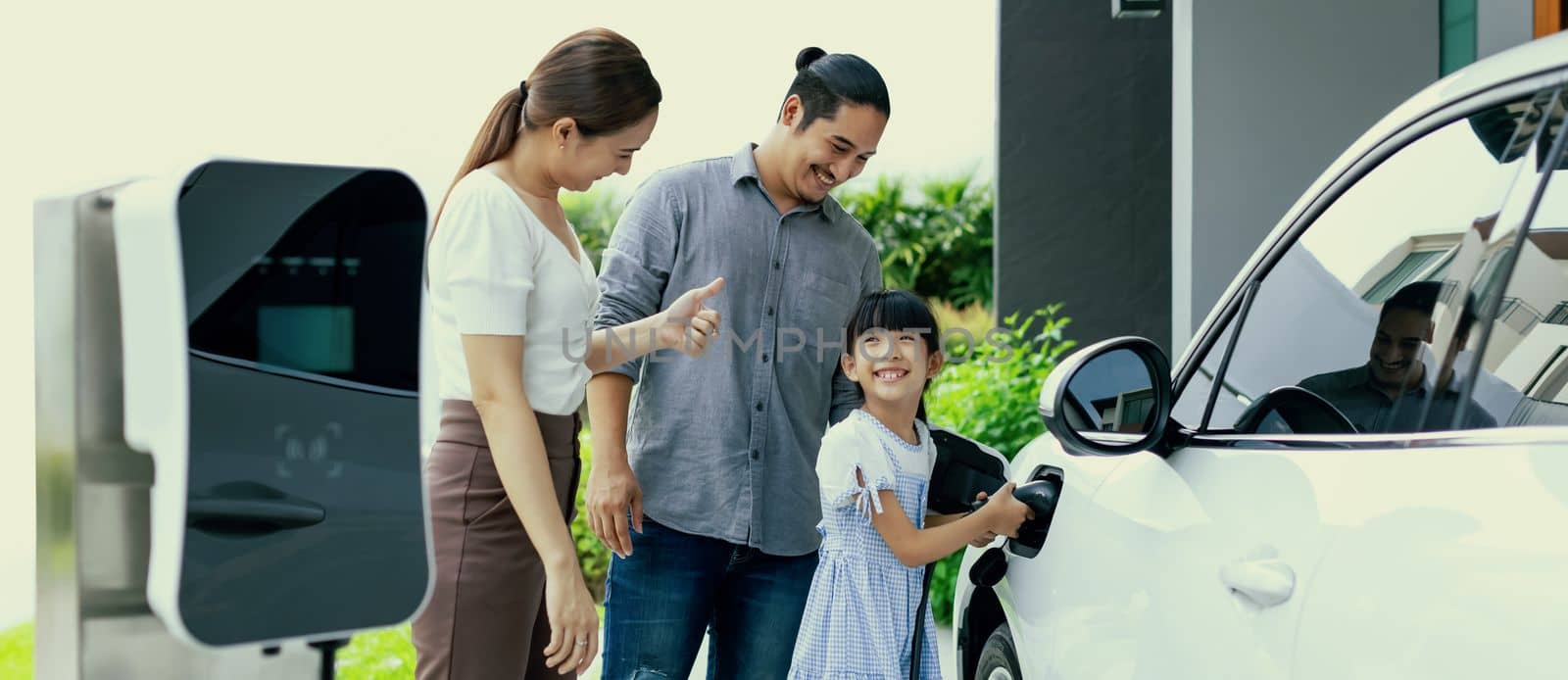 Progressive young parent teach daughter how to recharge or refuel EV car at home charging station. Green and clean energy from electric vehicle for healthy environment. Eco power from renewable source