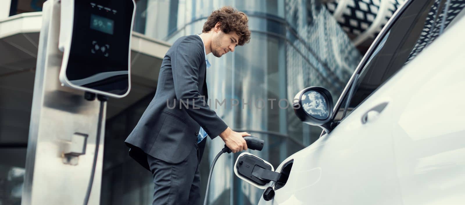 Below view closeup image of progressive black suit businessman recharge battery of his electric vehicle from public charging station. Renewable and alternative energy powered car concept.