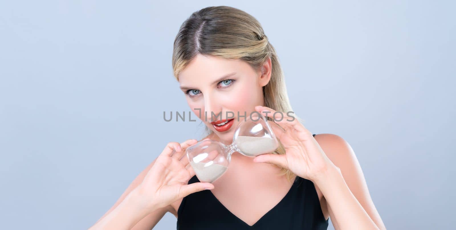 Alluring beauty model holding hourglass in forever young beauty concept of anti-aging skincare treatment for woman. Beautiful caucasian women portrait with perfect makeup in isolated background.