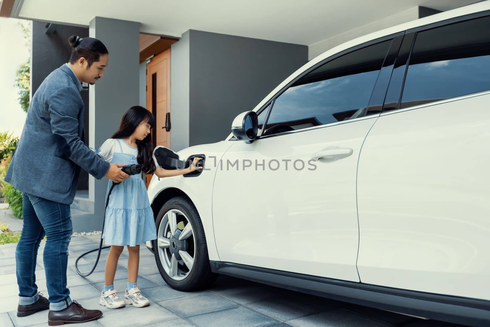 Progressive dad and daughter charging EV car from home charging station. by biancoblue