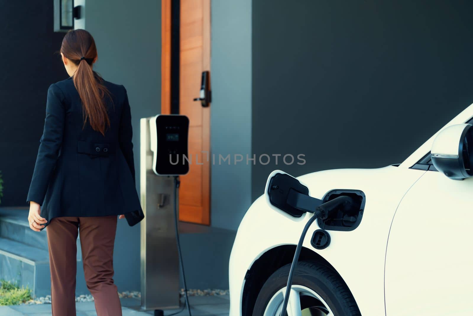 Progressive woman install cable plug to her electric car with home charging station. Concept of the use of electric vehicles in a progressive lifestyle contributes to clean environment.