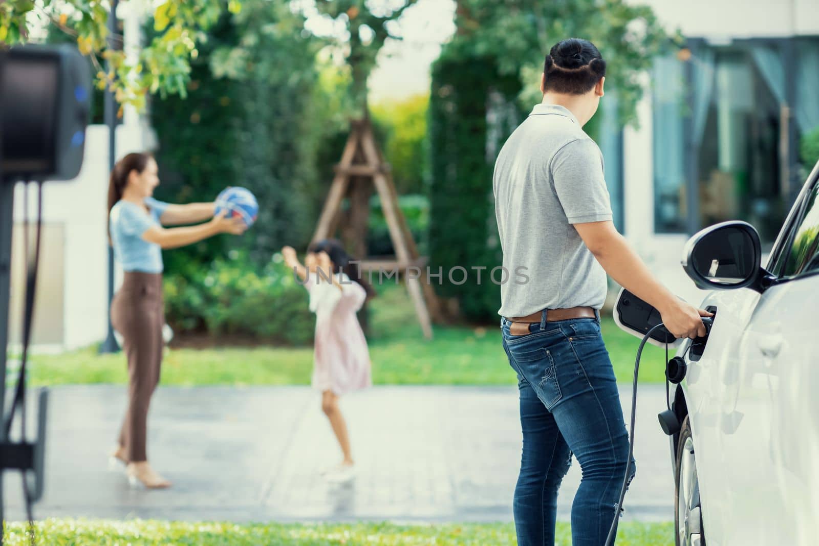 Focus image of progressive man charging electric car from home charging station with blur mother and daughter playing together in the background.