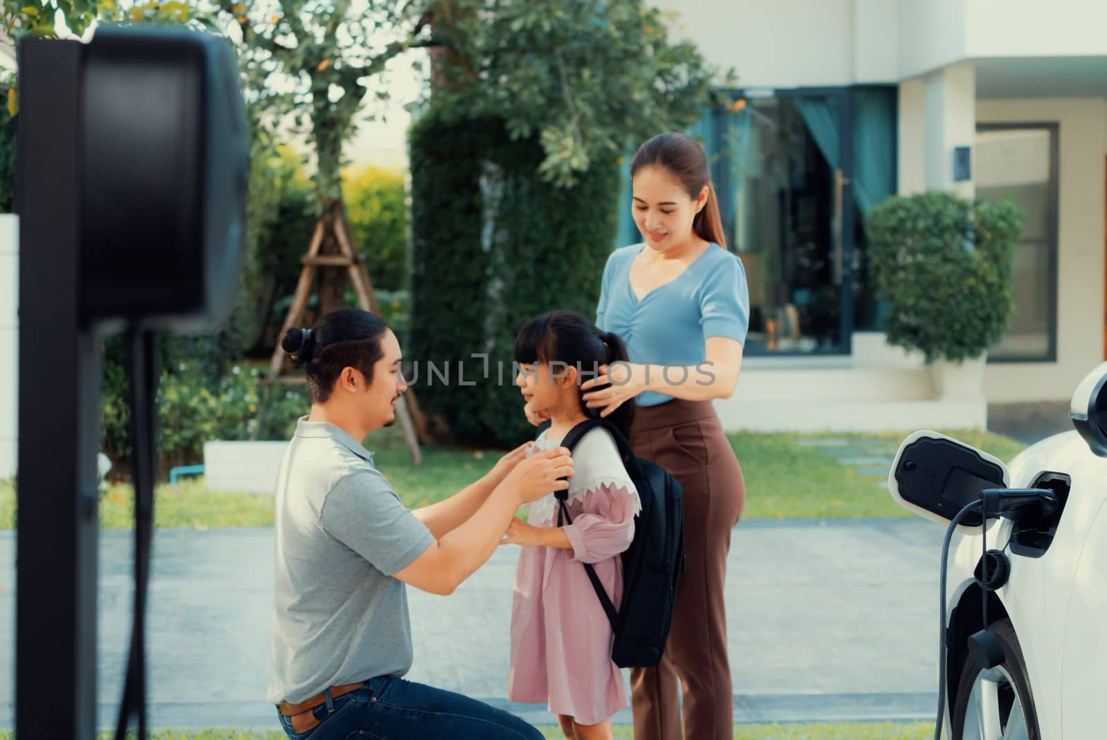 Progressive young parents and daughter with electric vehicle and home charging station. Green and clean energy from electric vehicles for healthy environment. Eco power from renewable source at home.