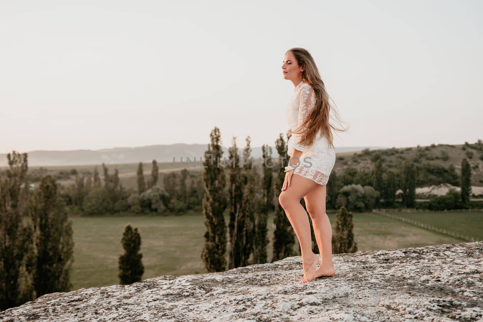 Romantic beautiful bride in white dress posing with sea and mountains in background. Stylish bride standing back on beautiful landscape of sea and mountains on sunset