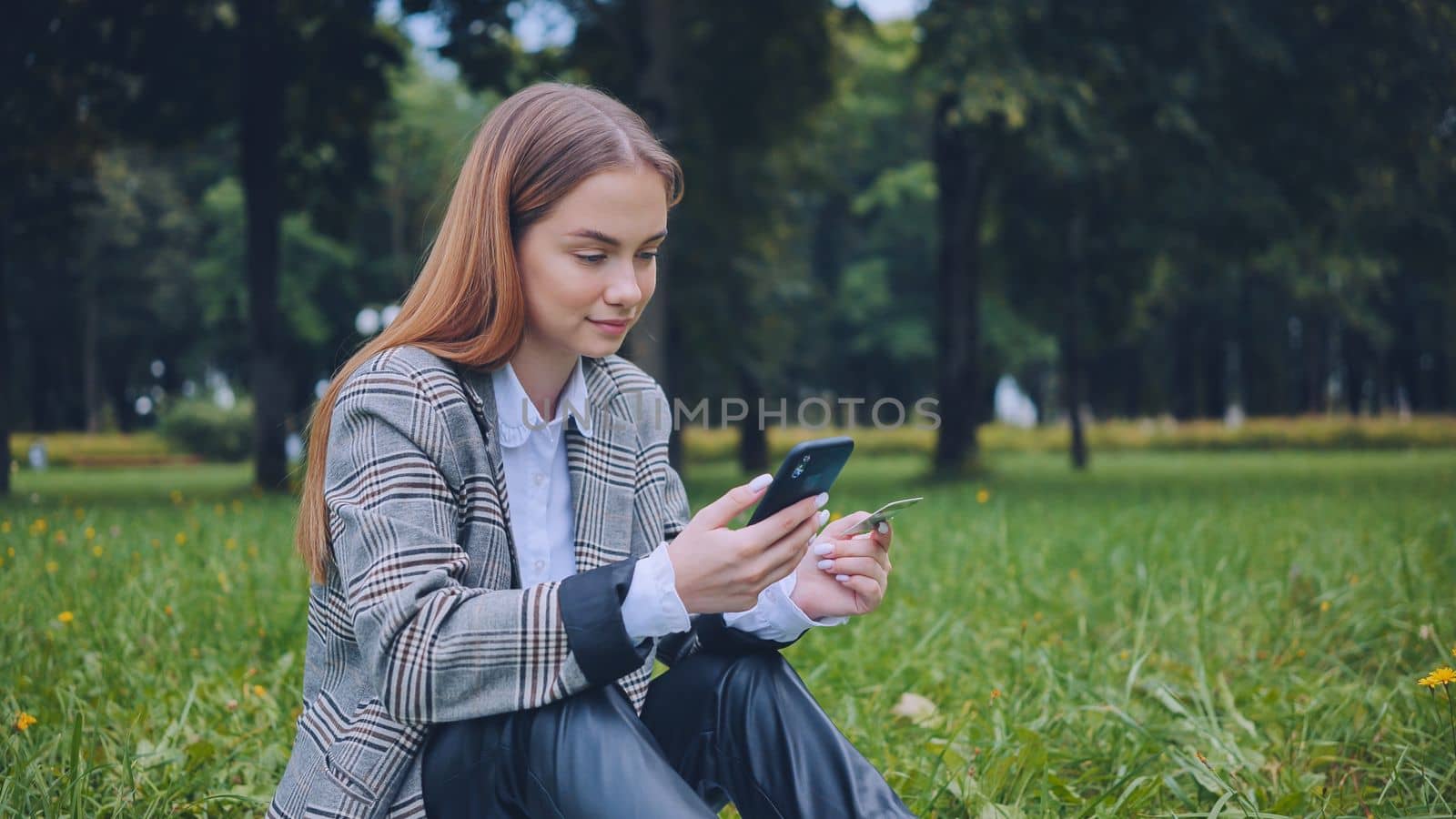 A young girl makes a purchase with her phone and bank card in the park, sitting on the grass
