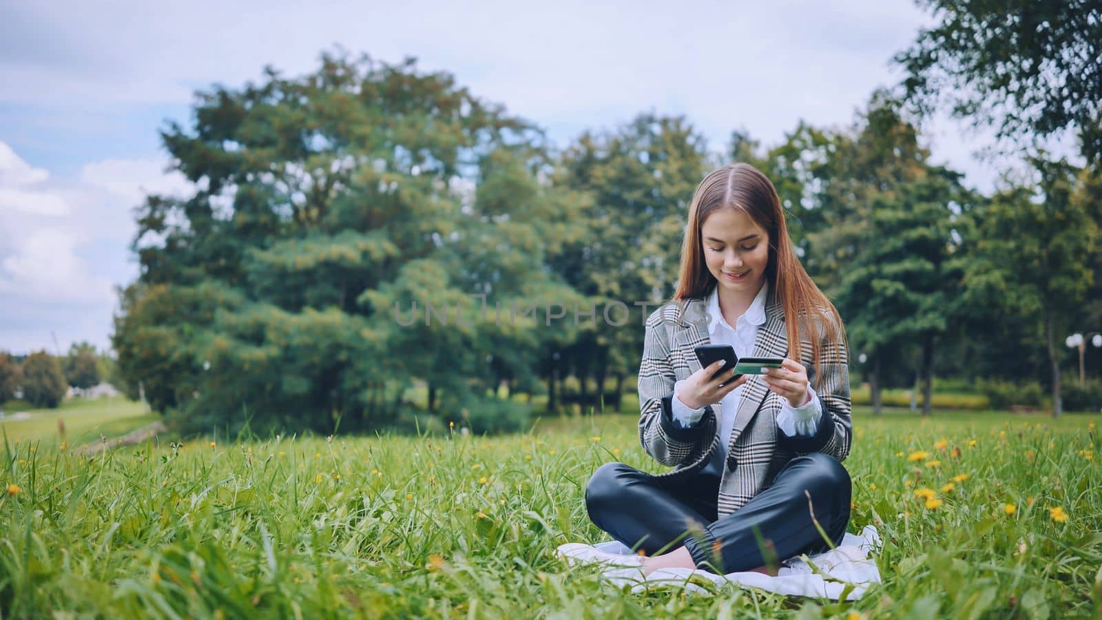 A young girl makes a purchase with her phone and bank card in the park, sitting on the grass