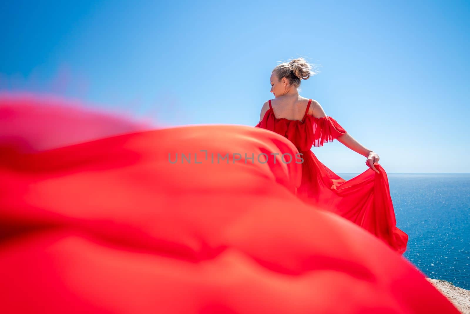 Blonde with long hair on a sunny seashore in a red flowing dress, back view, silk fabric waving in the wind. Against the backdrop of the blue sky and mountains on the seashore