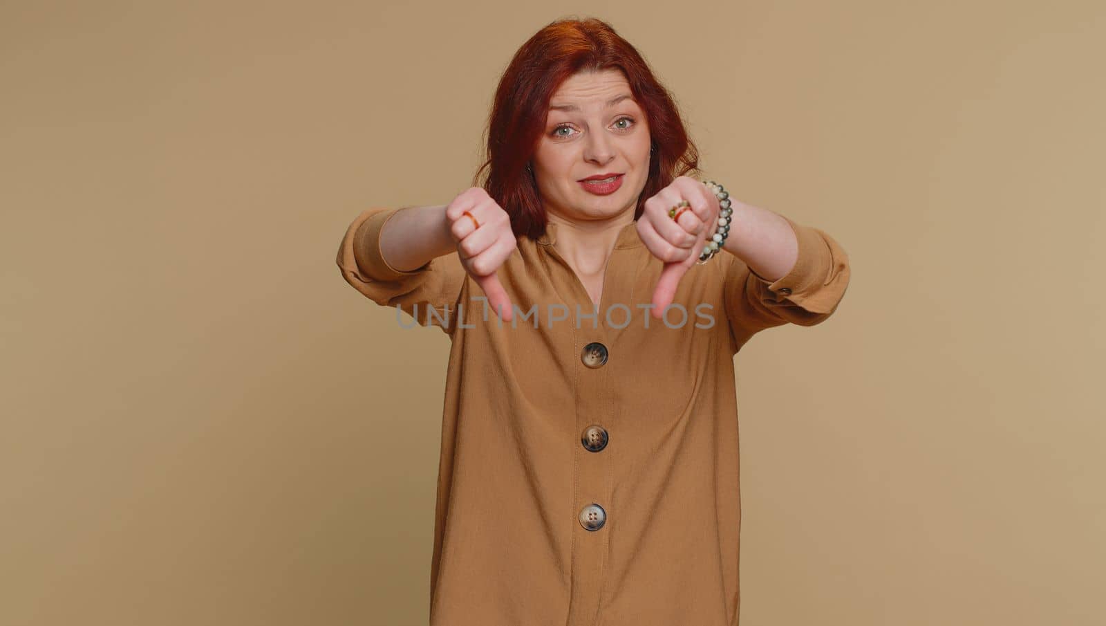 Dislike. Upset unhappy redhead woman in blouse showing thumbs down sign gesture, expressing discontent, disapproval, dissatisfied, dislike. Young adult girl. Indoor studio shot on beige background