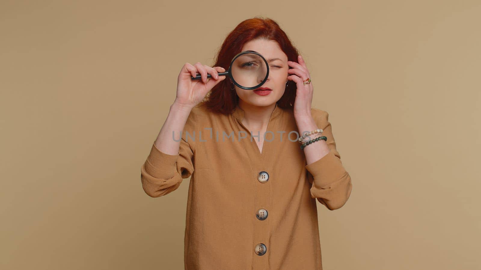 Investigator researcher scientist woman holding magnifying glass near face, looking into camera with big zoomed funny eyes, searching, analysing. Young redhead girl isolated on beige studio background