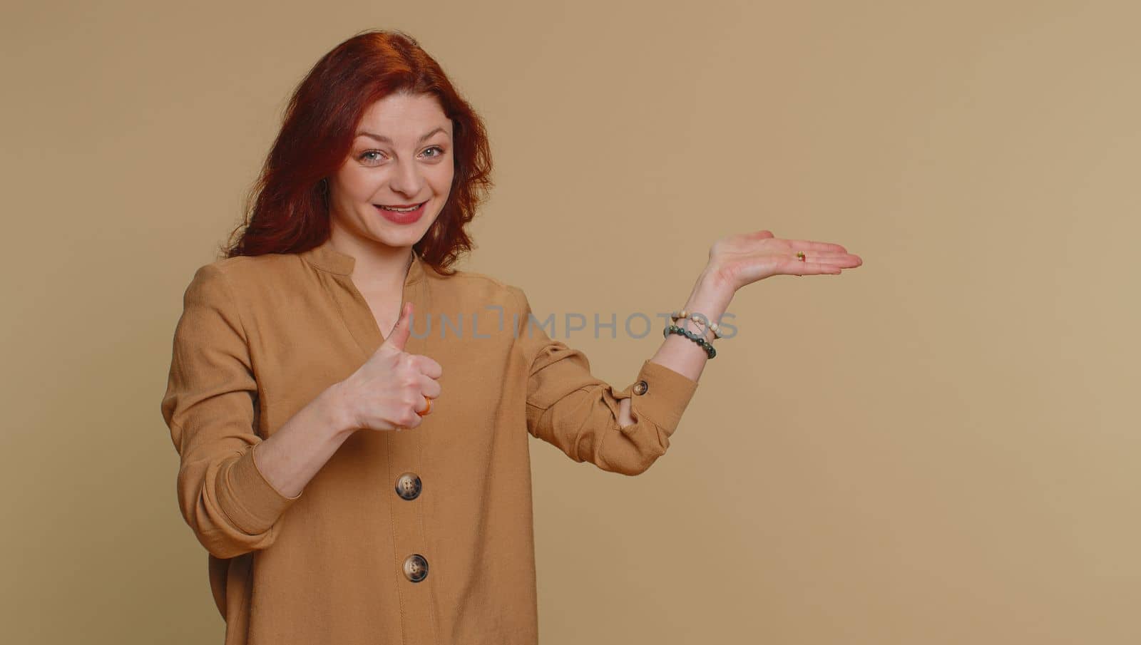 Lovely woman in blouse showing thumbs up and pointing empty place, advertising area for commercial text, copy space for goods promotion. Young redhead girl. Indoor studio shot on beige wall background