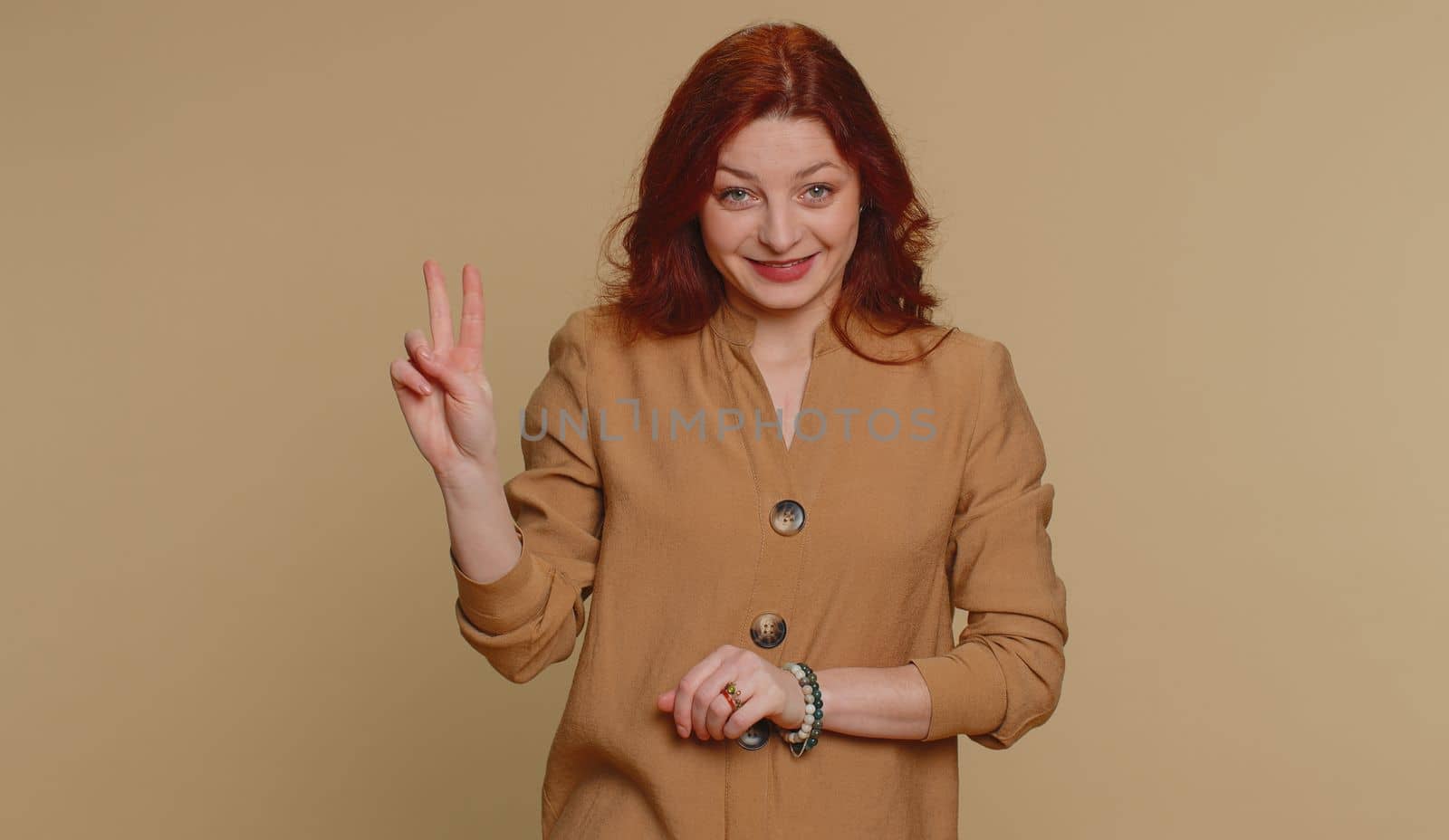Hipster redhead woman in blouse showing victory v sign, hoping for success and win, doing peace gesture, smiling with kind optimistic expression. Young adult girl isolated on beige studio background