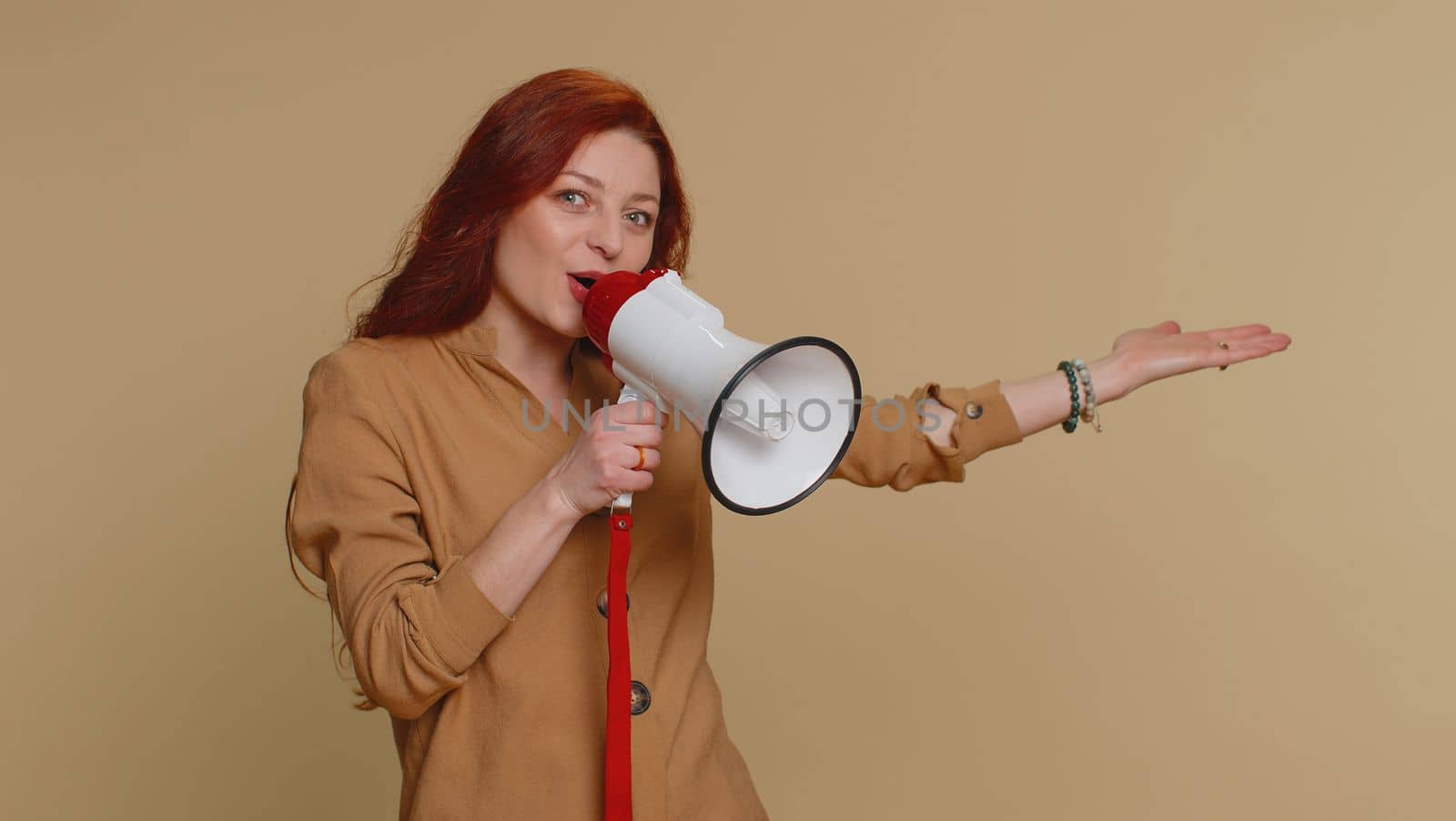 Redhead woman talking with megaphone, proclaiming news, loudly announcing advertisement, warning using loudspeaker to shout speech, pointing empty place. Copy-space. Young girl on beige background