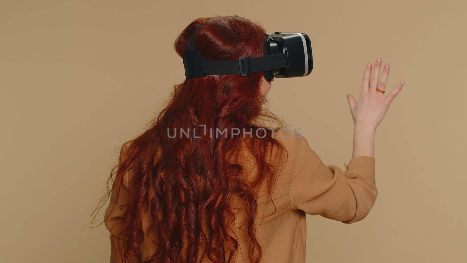 Woman using headset helmet to play simulation game app online. Watching virtual reality 3D 360 video content. Girl in VR goggles isolated on studio beige background. Futuristic technology. Rear view