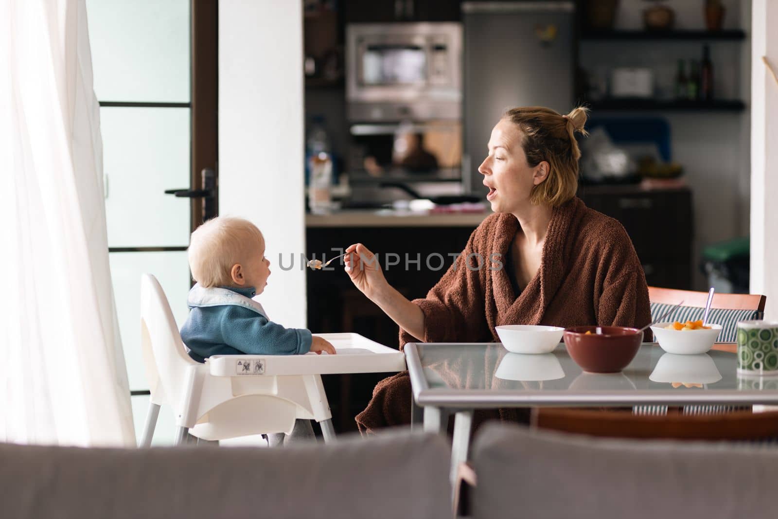 Mother wearing bathrope spoon feeding her infant baby boy child sitting in high chair at the dining table in kitchen at home in the morning.