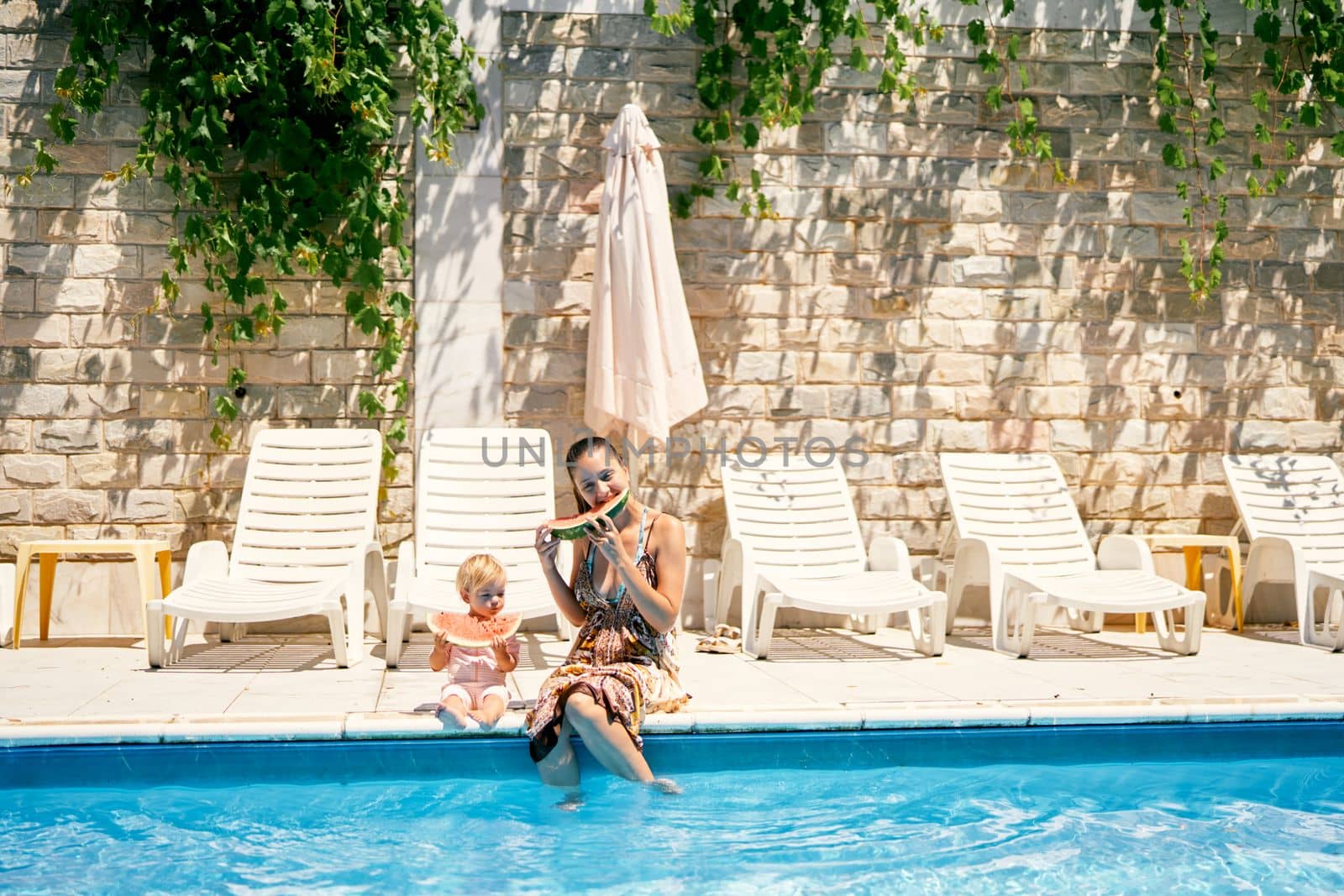 Mom and little girl eat watermelon sitting by the pool. High quality photo