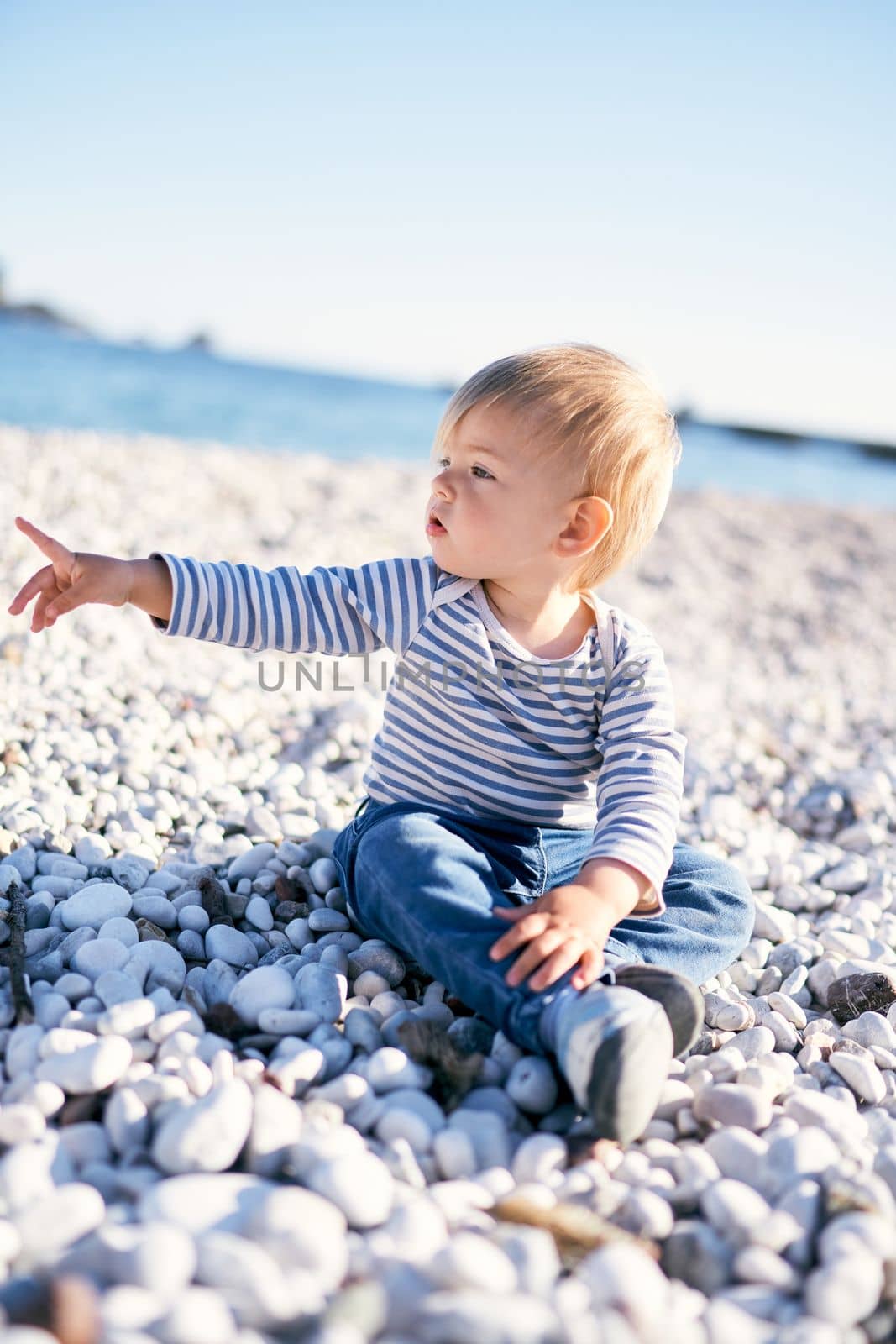 Kid sits on a pebble beach by the sea and points his finger into the distance. High quality photo