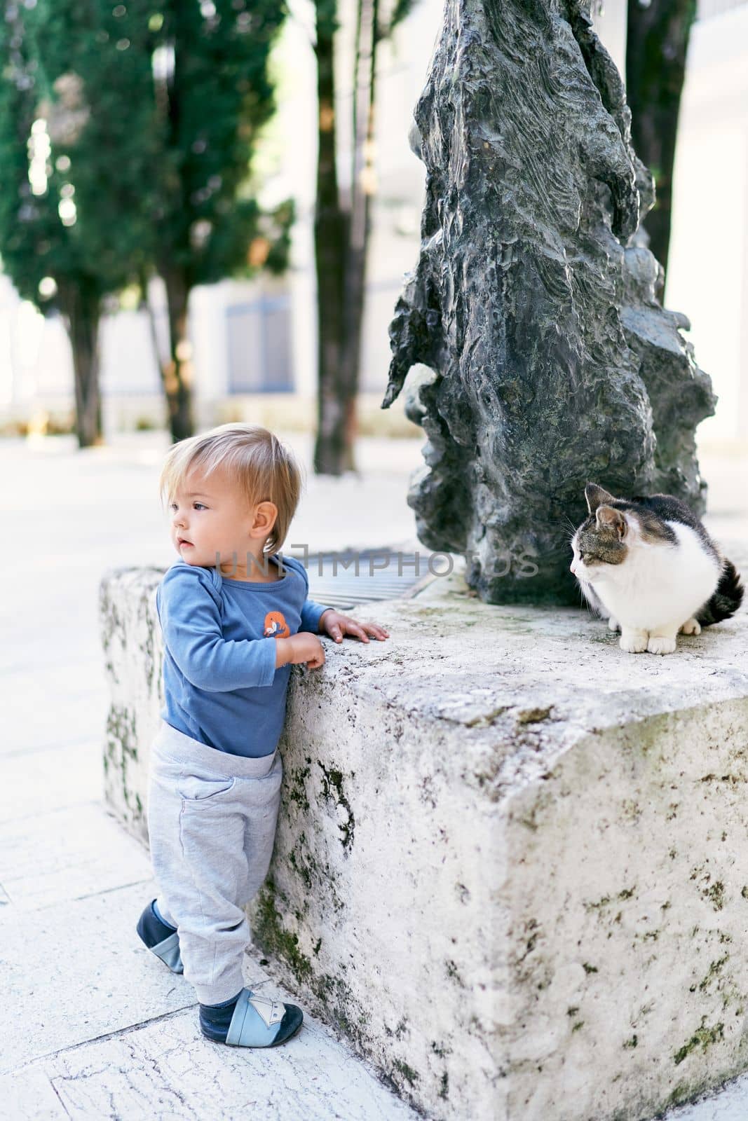 Kid stands, turned sideways, near the sculpture on which the cat sits by Nadtochiy