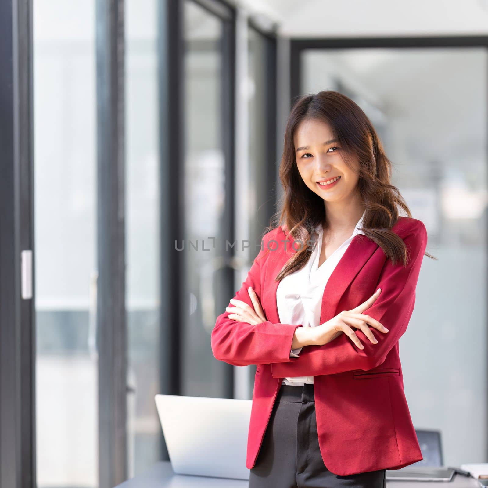 Businesswoman entrepreneur or an office worker stands with crossed arms near a desk in a office, looking at the camera and smiling.