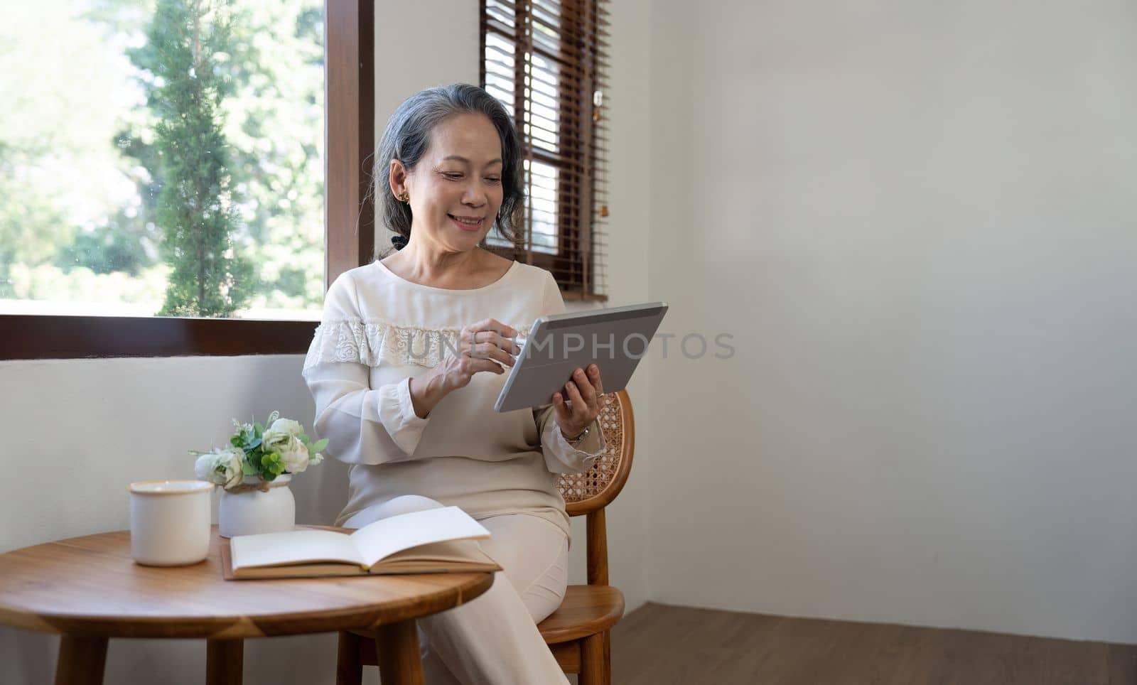 Happy aged Asian old woman sitting by the window and using tablet by nateemee