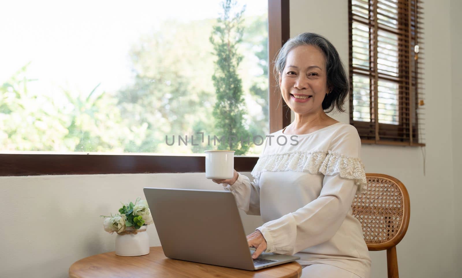 Happy aged Asian old woman working , sipping coffee while using laptop.