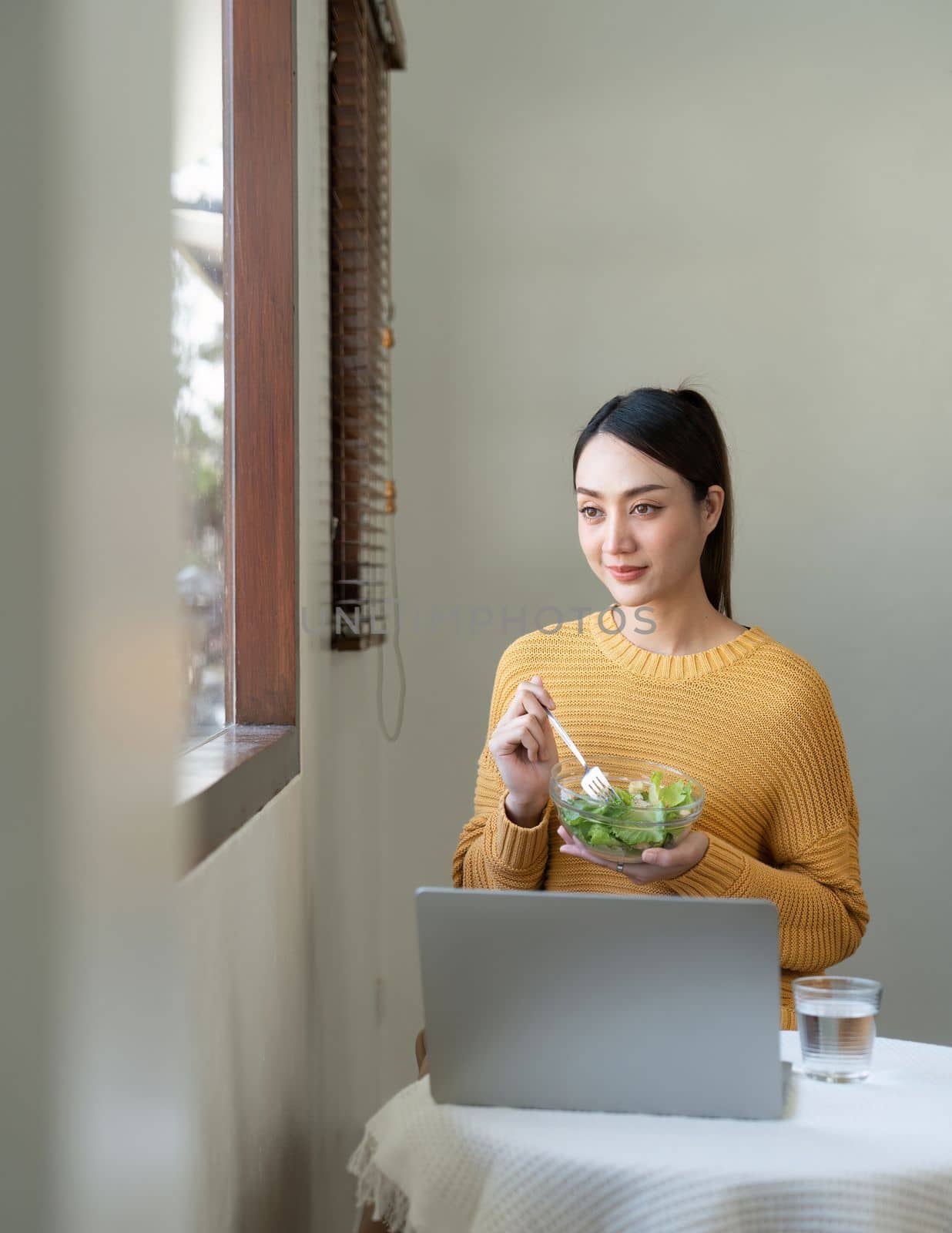 Happy Asian woman eating healthy salad vegetables mix while using her laptop in the living room. work from home.