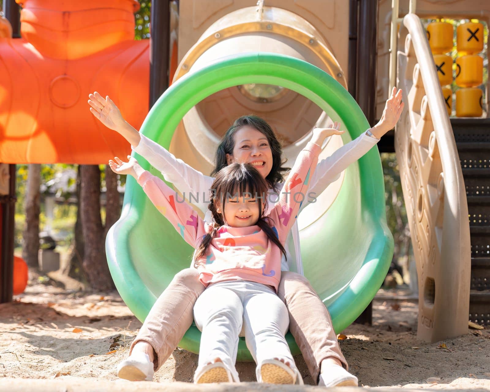 Happy Asian little girl playing on slider, enjoys playing on playground with her grandmother.