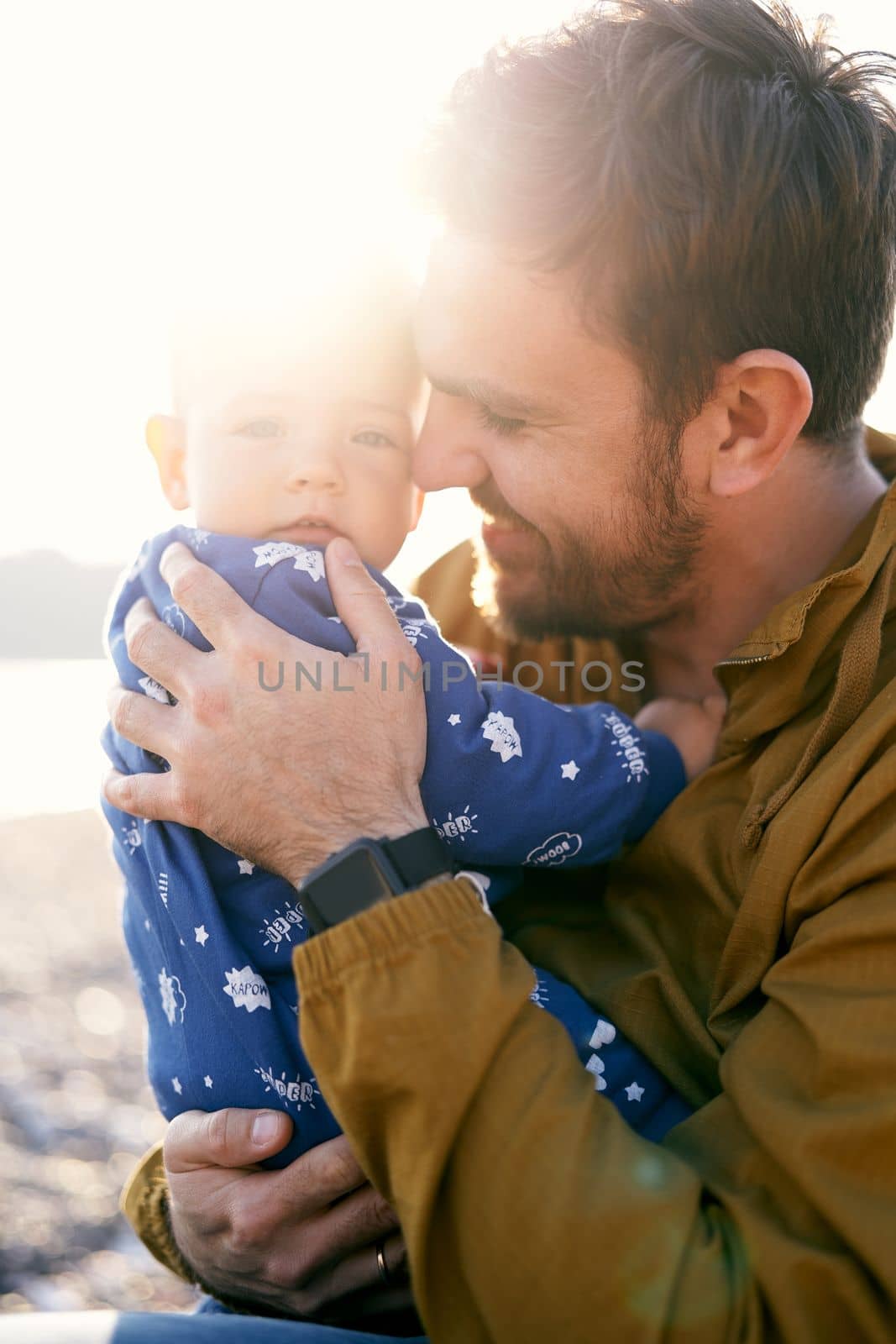 Smiling dad in a brown jacket sits and holds in his arms a small child in a blue overalls. Sun shines brightly. Close-up by Nadtochiy