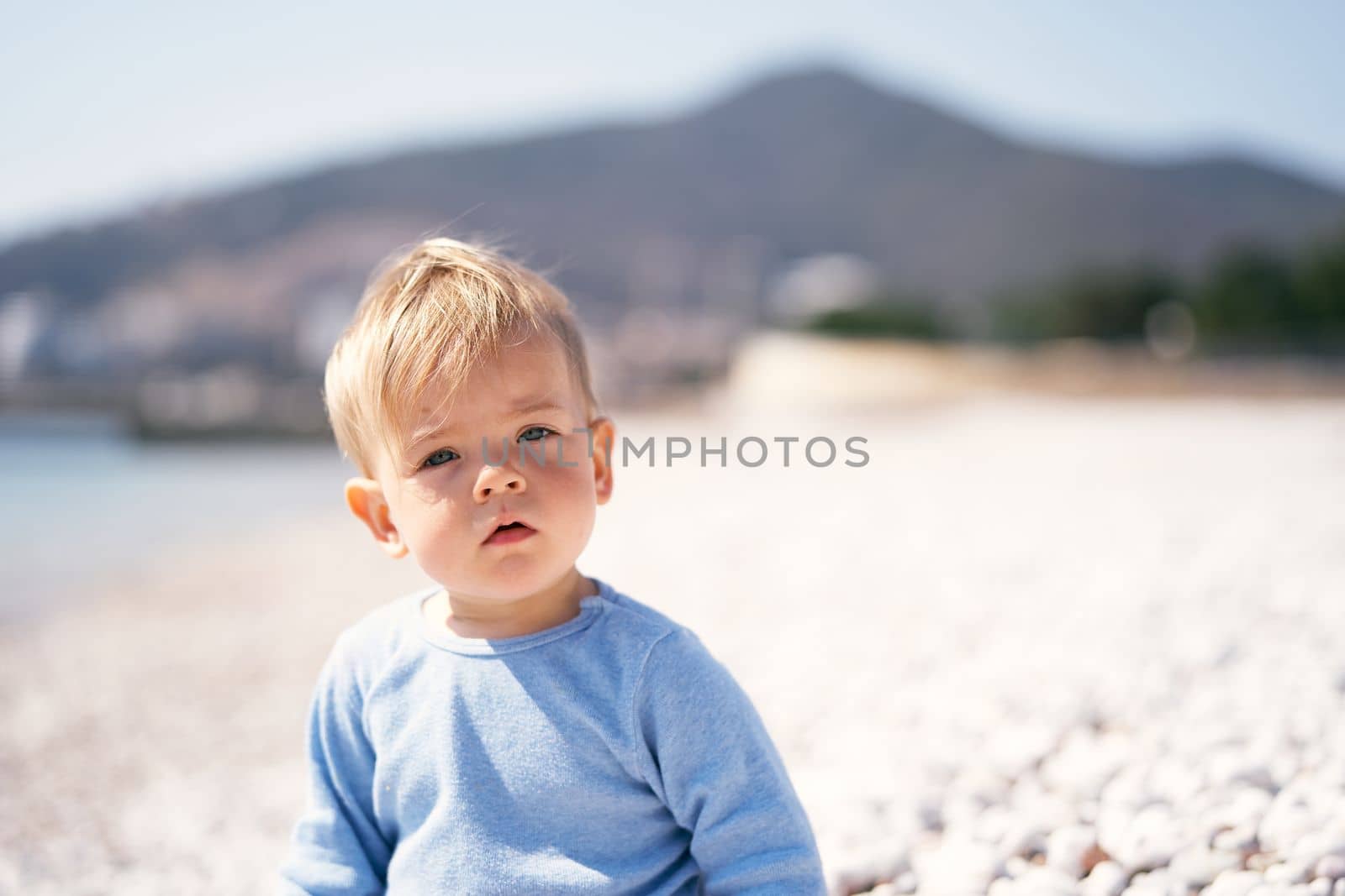 Serious child sits on a pebble beach by the sea against the backdrop of mountains. Close-up. Portrait by Nadtochiy